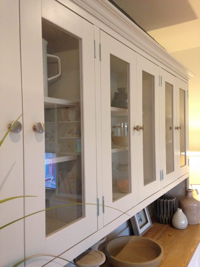 Angled view along the glazed rack from the side, showing the six glass doors with brushed nickel knobs, the shelves of jars and jugs inside and the props on the pine top below