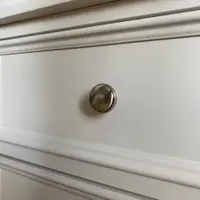 Detail of one brushed nickel knob on a cream painted drawer front, showing the fine beaded moulding around the panel and the moulded lip of the cabinet top above