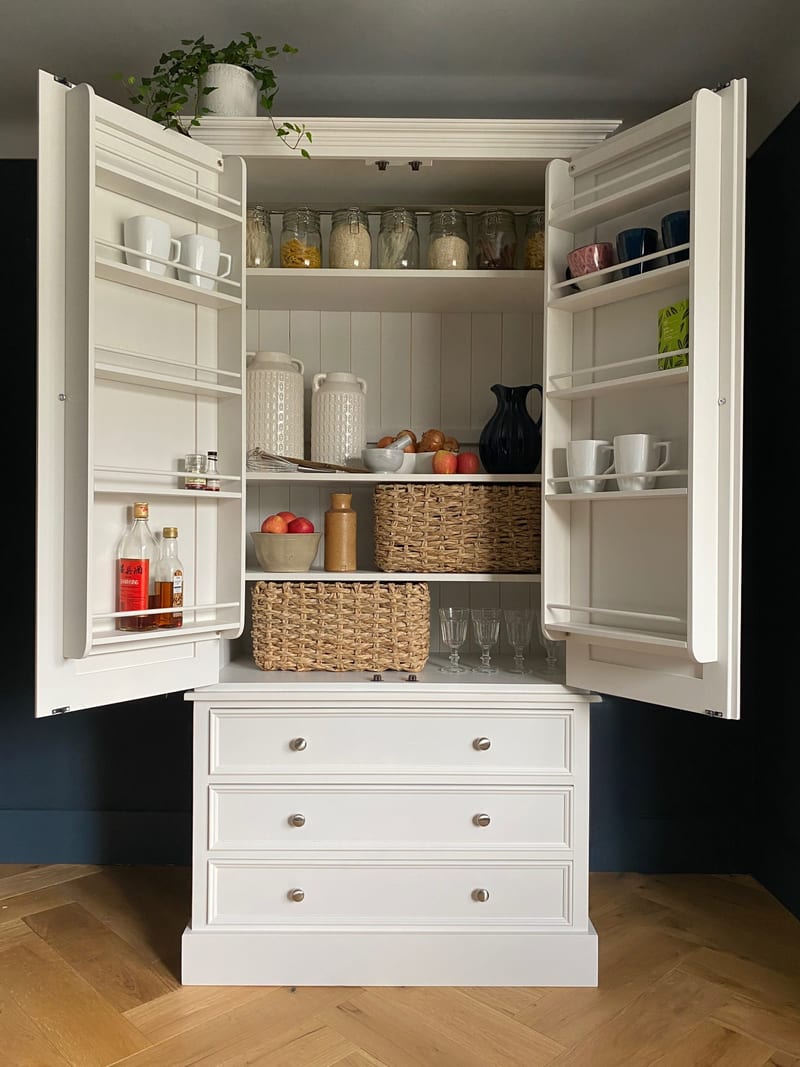 Full front view of the cream larder cabinet with both doors open, showing the fitted door racks, three interior shelves of jars and baskets and the three drawers below against a navy wall