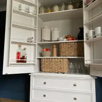 Angled view into the open larder, showing the left door rack of bottles and mugs, the tongue-and-groove back, shelves of jars, baskets and glasses and the three drawers beneath