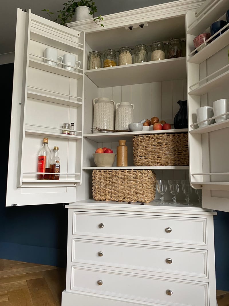 Angled view into the open larder, showing the left door rack of bottles and mugs, the tongue-and-groove back, shelves of jars, baskets and glasses and the three drawers beneath