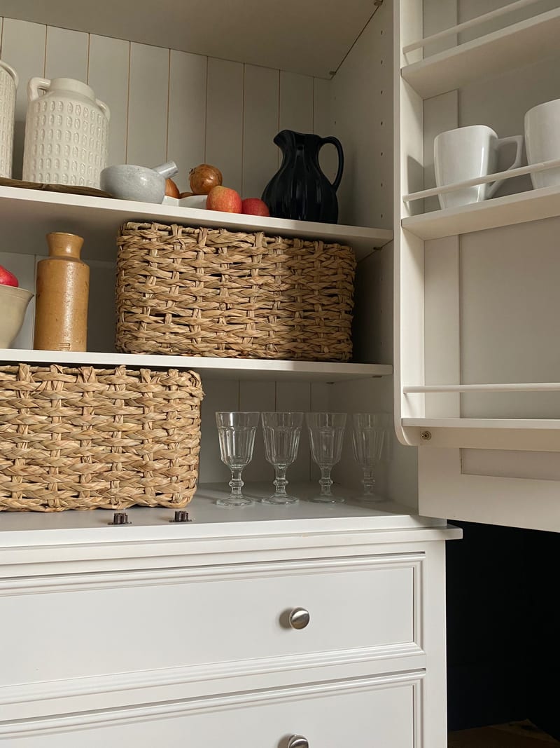 Close view of the larder shelves, showing the tongue-and-groove back board, two woven seagrass baskets, a black jug, cut glasses and the mug rack fixed to the open door