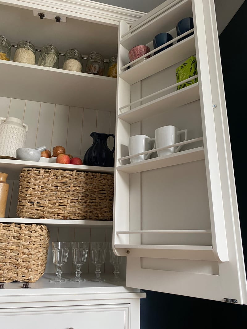 Close view of the open right-hand door with its fitted rack, showing the retaining rails holding mugs and bowls and the interior shelves of jars and baskets alongside