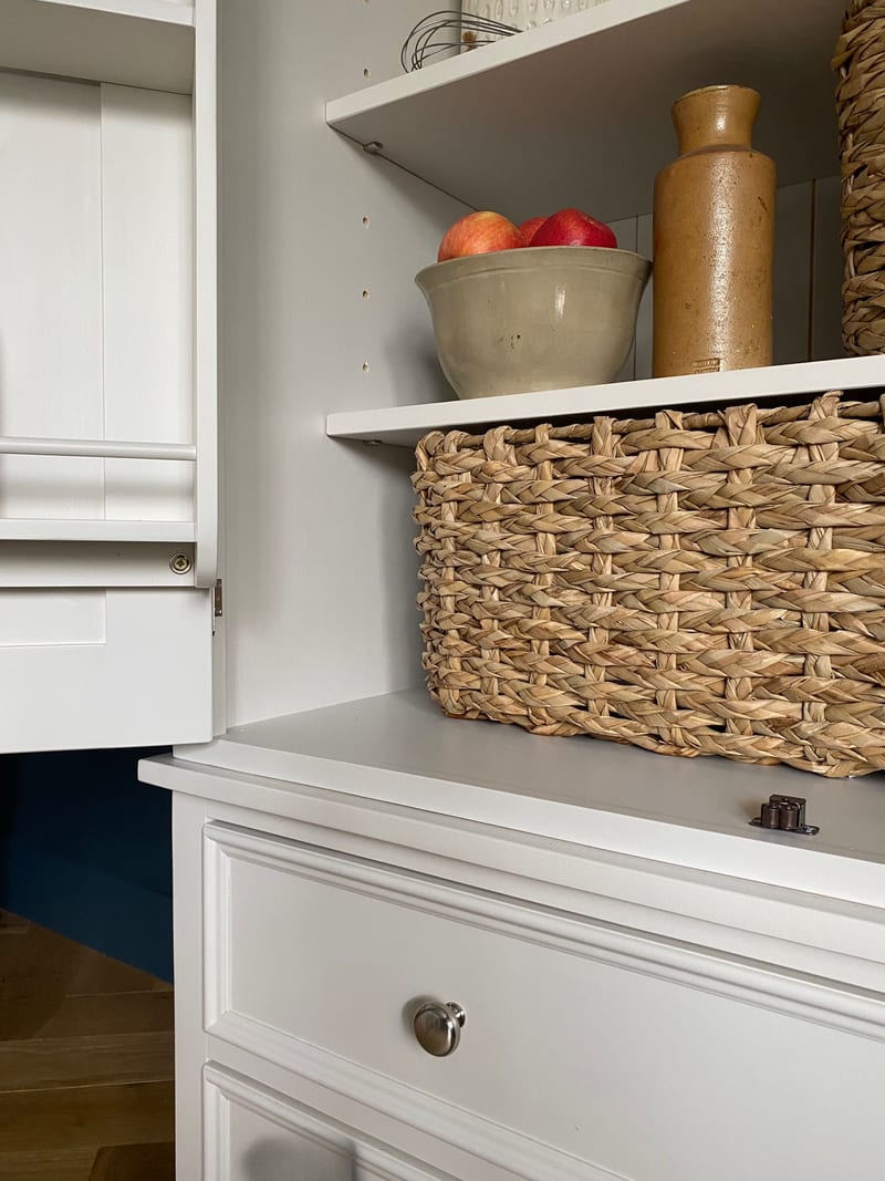 Close view inside the larder, showing an adjustable shelf with a stoneware bowl of apples and a tall jar, a seagrass basket on the base top and a drawer front with a nickel knob