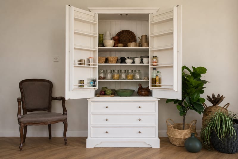White painted larder cupboard with both doors opened to show spice racks on the doors and shelves of jars, bowls and baskets inside, above three deep drawers in the base