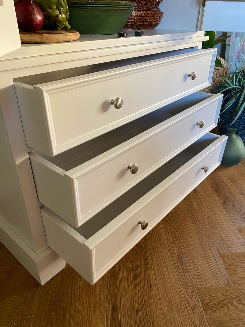 Angled close view of the three base drawers of the white larder cabinet pulled out in a staircase, showing the moulded fronts, brushed nickel knobs and the oak herringbone floor beneath