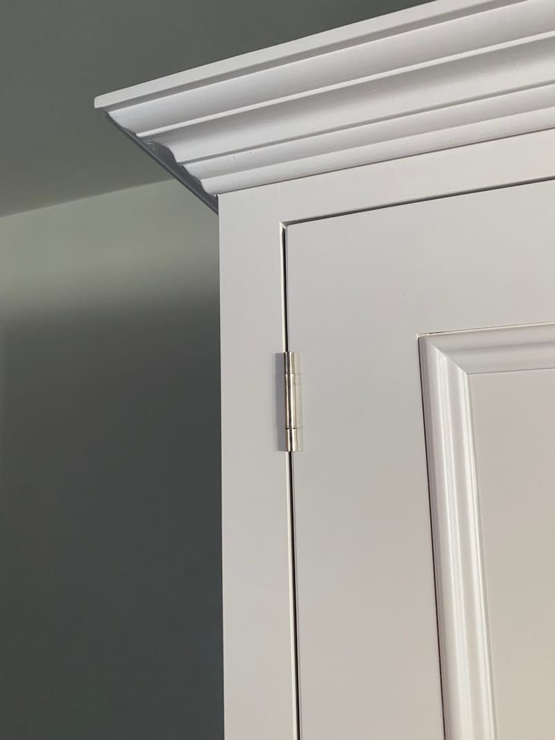 Detail of the top corner of the white larder cabinet, showing the stepped moulded cornice, panelled door edge and polished nickel butt hinge against a dark sage green wall