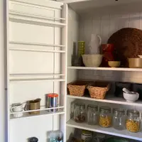 Close view inside the open left-hand door of the white larder cabinet, showing the door spice racks, tongue-and-groove back and shelves of jugs, wicker baskets and glass storage jars