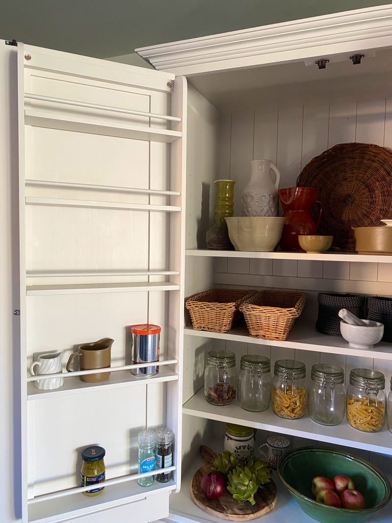 Close view inside the open left-hand door of the white larder cabinet, showing the door spice racks, tongue-and-groove back and shelves of jugs, wicker baskets and glass storage jars