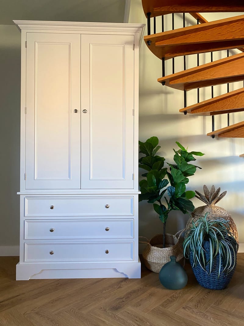 Full view of the white larder cabinet with both doors closed, two panelled doors over three drawers with brushed nickel knobs, standing beside an oak spiral staircase and potted plants