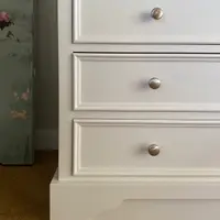 Close view of the three drawer fronts and shaped plinth of the white larder cabinet, showing the moulded drawer edges, brushed nickel knobs and the oak floor below