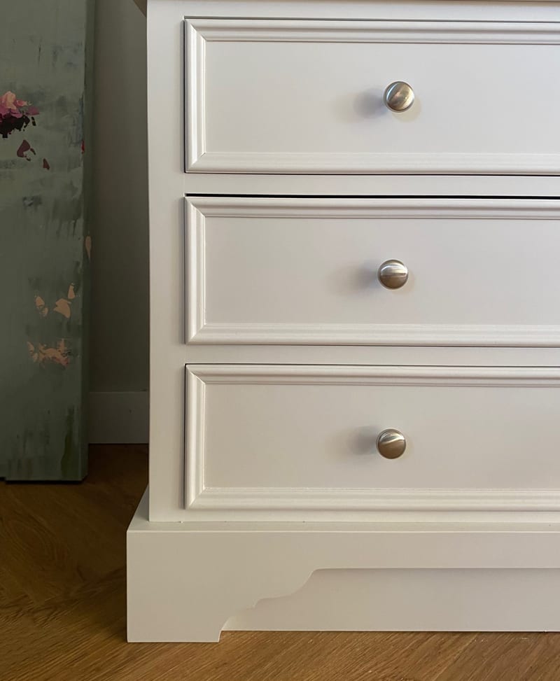 Close view of the three drawer fronts and shaped plinth of the white larder cabinet, showing the moulded drawer edges, brushed nickel knobs and the oak floor below