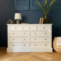 Cream painted large chest with three short drawers above three pairs of long drawers on a deep plinth, against a dark navy wall on herringbone oak flooring with a lamp and books on top