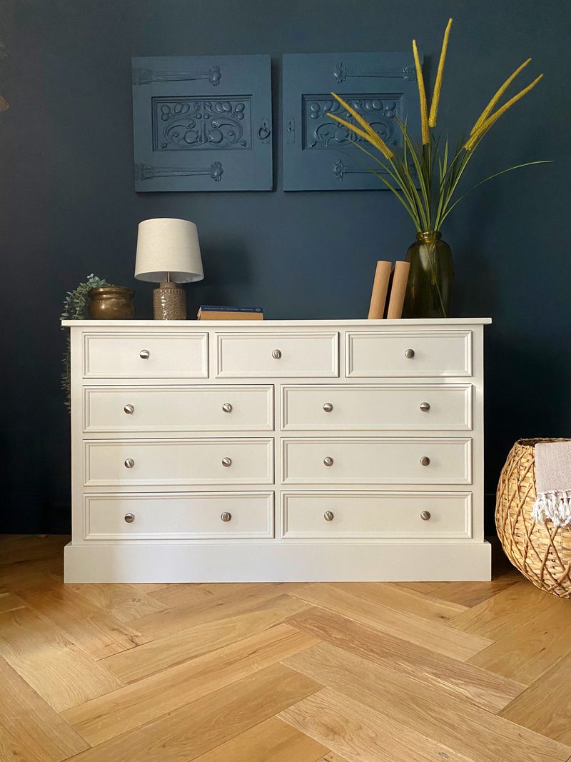 Cream painted large chest with three short drawers above three pairs of long drawers on a deep plinth, against a dark navy wall on herringbone oak flooring with a lamp and books on top