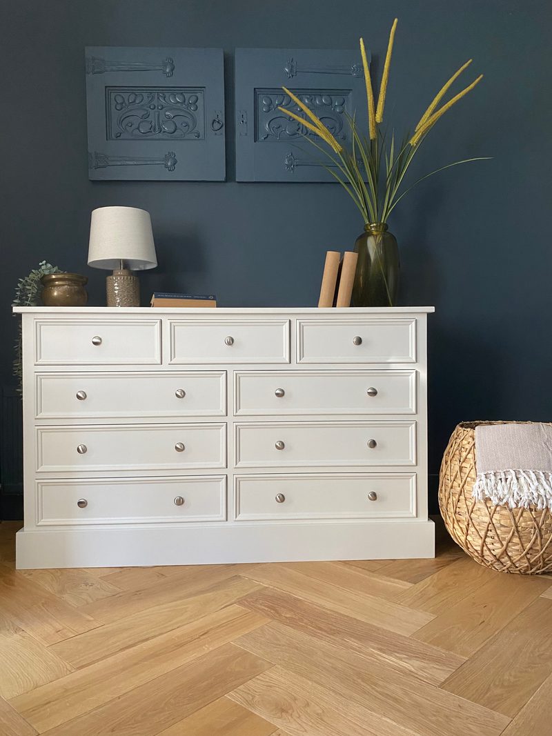 Close front view of the off-white chest of drawers styled with a table lamp, brass pot, books and a vase of dried grasses, with a fringed woven basket beside it on herringbone parquet