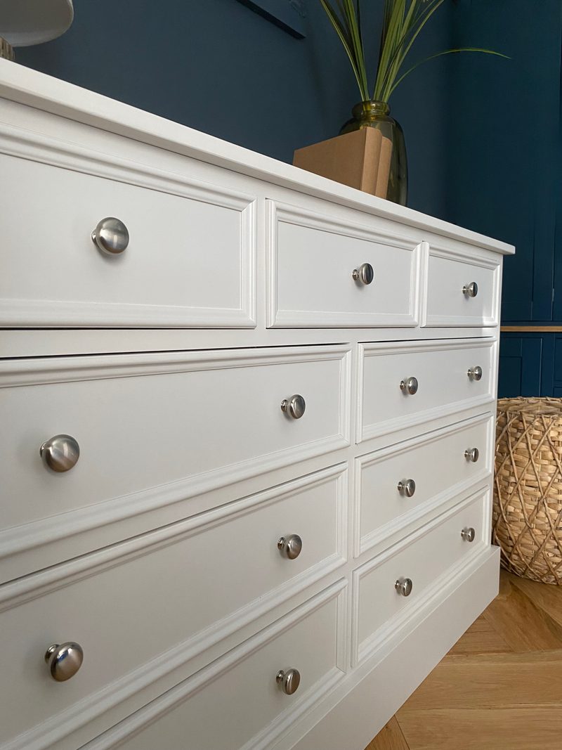 Low angled view along the off-white drawer fronts, showing the moulded panel edges and rows of brushed nickel knobs receding towards the deep navy wall and woven basket