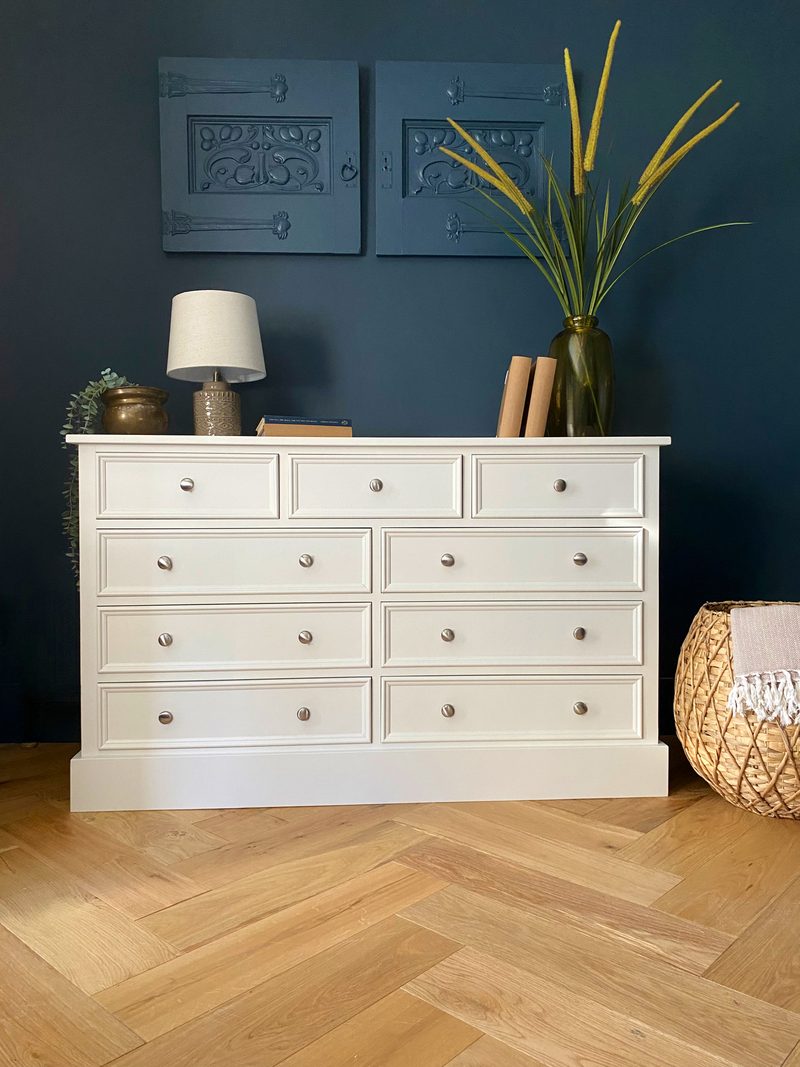 Full front view of the off-white nine-drawer chest, three short drawers above three rows of paired wide drawers, all with brushed nickel knobs, on light oak herringbone parquet