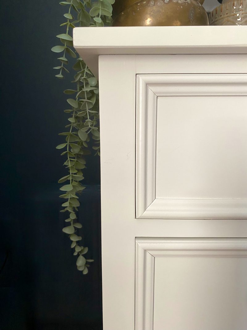Close view of the top left corner of the off-white chest, showing the overhanging top edge, moulded drawer fronts and a trailing eucalyptus stem against the dark navy wall