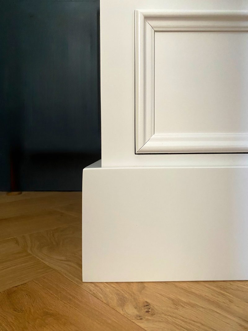 Detail of the bottom left corner of the off-white chest, showing the square plinth, moulded drawer front and shadow gap above the light oak herringbone parquet