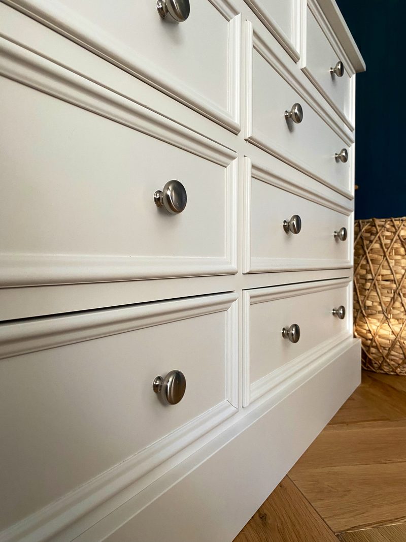 Close angled view of the drawer fronts of the off-white chest from below, showing the moulded borders, brushed nickel knobs and plinth beside a woven basket