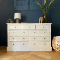 Cream painted nine drawer chest on a deep plinth against a dark navy wall, with a lamp, books and dried grasses on top and herringbone oak flooring below