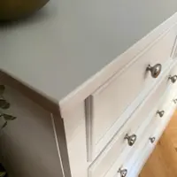 Overhead close view across the painted top of the off-white chest, showing the moulded top edge, side panel and upper drawer fronts with brushed nickel knobs, beside a brass vessel and an open book
