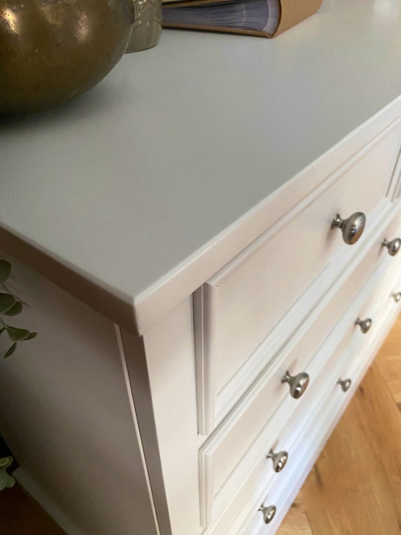 Overhead close view across the painted top of the off-white chest, showing the moulded top edge, side panel and upper drawer fronts with brushed nickel knobs, beside a brass vessel and an open book