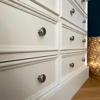 Low close view along the paired drawer fronts of the off-white chest, showing the moulded borders, brushed nickel knobs and the plinth running towards a woven basket on oak parquet