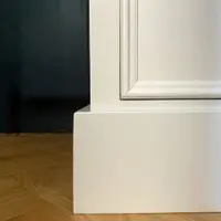 Corner detail of the off-white painted base, showing the square plinth, the moulded lower drawer front and the light oak herringbone parquet running up to a deep navy wall