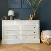 Front view of the off-white chest of drawers dressed with a linen shade lamp, brass pot, books and dried grasses, with a fringed woven basket alongside on parquet flooring