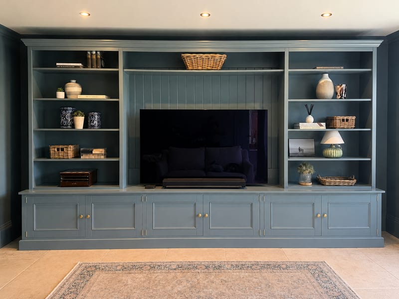 Blue grey painted 12ft Newton media wall with a large television in the centre, open shelving either side holding books, baskets and ceramics, and cupboards running the full width below