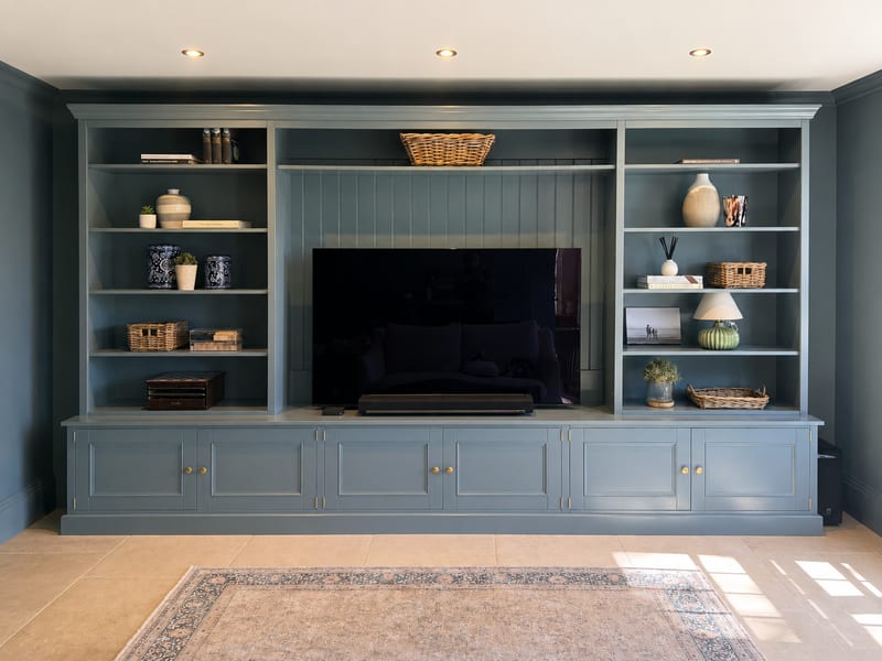 Straight-on view of the blue-grey painted media wall unit, open shelving either side of the tongue-and-groove television recess, brass-knobbed base cupboards and sunlight across the stone floor tiles