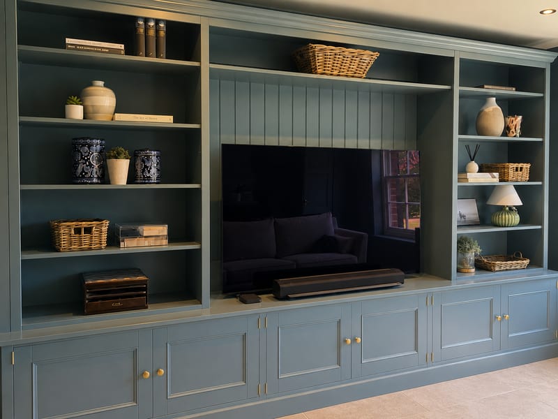 Angled view along the left-hand bookcase and central recess, showing shelves styled with stoneware vases, wicker baskets and books above the panelled cupboard doors with brass knobs