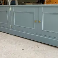 The painted base carcass photographed in the workshop before installation, showing the run of panelled cupboard doors, brass knobs and the moulded plinth on a concrete floor