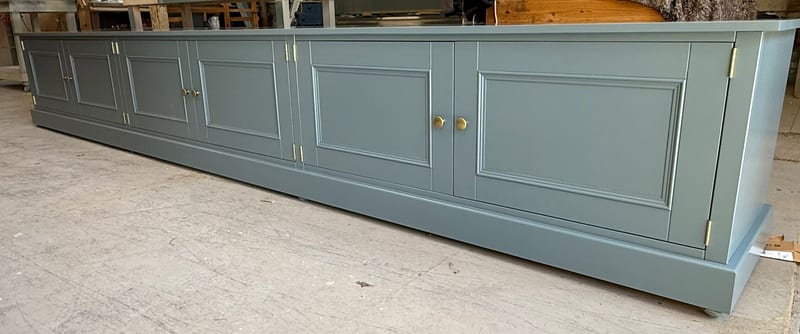 The painted base carcass photographed in the workshop before installation, showing the run of panelled cupboard doors, brass knobs and the moulded plinth on a concrete floor