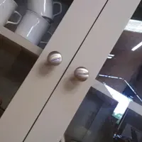 Close angled view of two glazed doors on the painted top section, the brushed nickel knobs set on the meeting stiles with speckled mugs and storage jars behind the glass