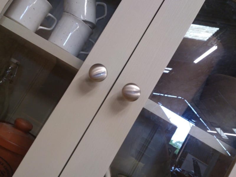 Close angled view of two glazed doors on the painted top section, the brushed nickel knobs set on the meeting stiles with speckled mugs and storage jars behind the glass