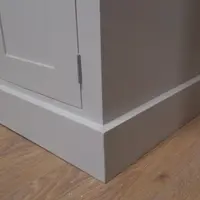 Detail of the corner of the grey painted base and its stepped plinth, showing the door edge, slim metal hinge and the oak-effect laminate flooring beneath