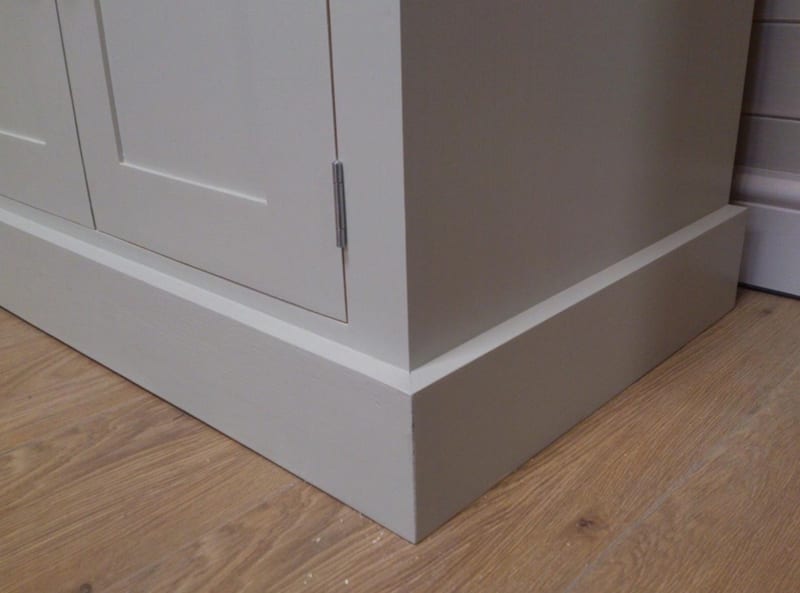 Detail of the corner of the grey painted base and its stepped plinth, showing the door edge, slim metal hinge and the oak-effect laminate flooring beneath