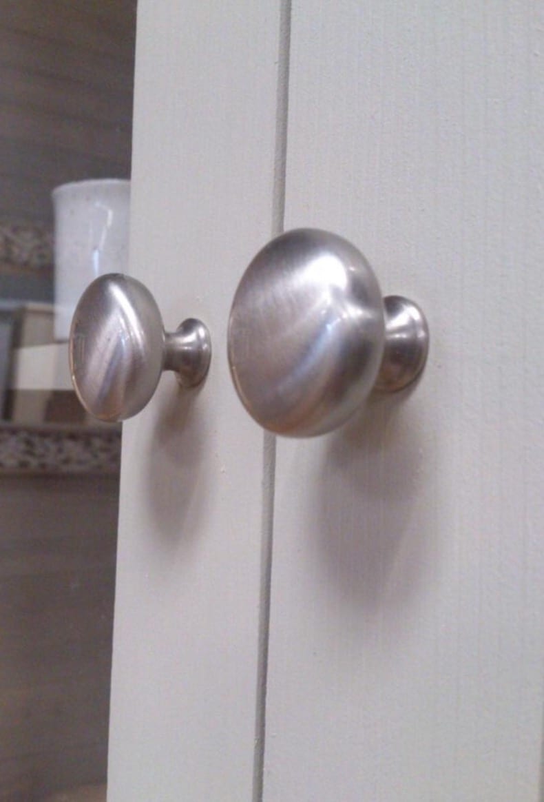 Macro detail of two brushed nickel oval knobs on the pale painted doors, with a white storage pot and a woven basket blurred in the cupboard behind the glass