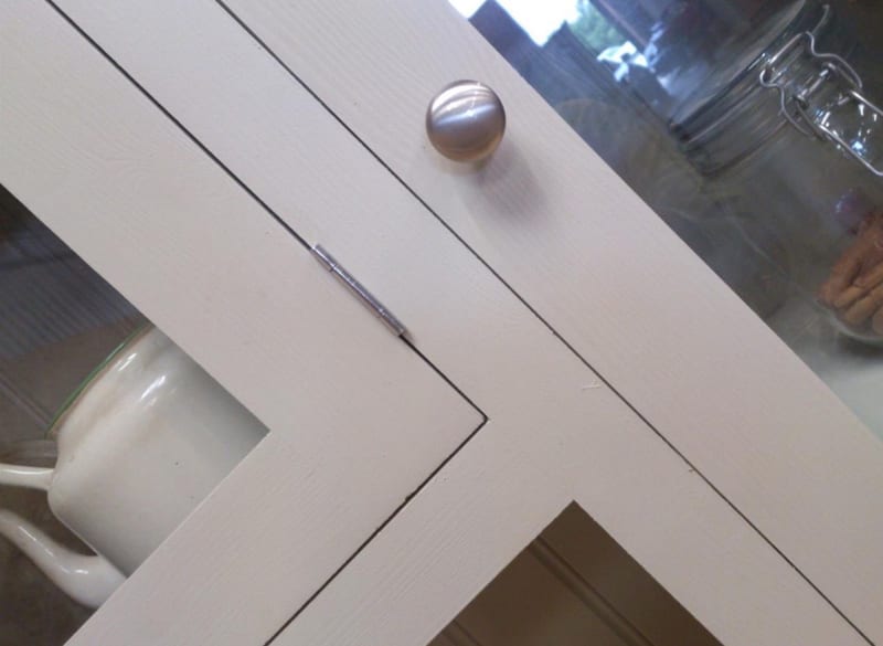 Close diagonal detail of a glazed door frame, showing the brushed nickel knob, slim steel hinge, painted astragal and the mug and clip-top jar visible through the glass