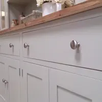 Low angled view along the base, showing three drawers over panelled cupboard doors with brushed nickel knobs beneath the waxed pine top, with a tripod floor lamp behind