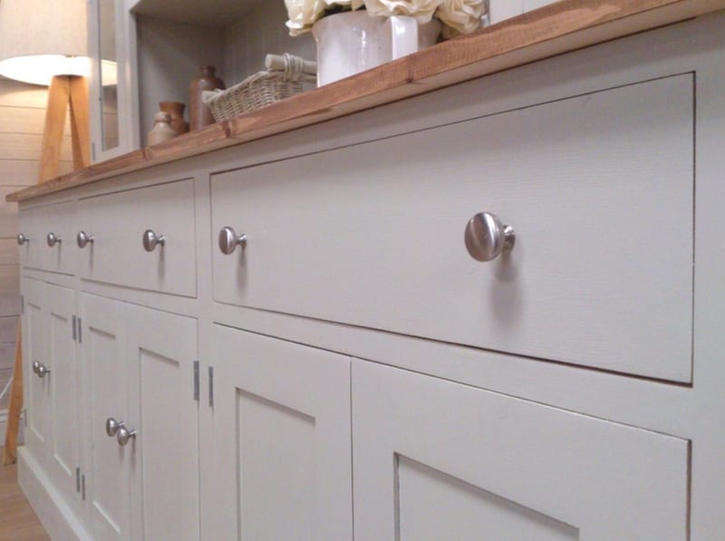 Low angled view along the base, showing three drawers over panelled cupboard doors with brushed nickel knobs beneath the waxed pine top, with a tripod floor lamp behind