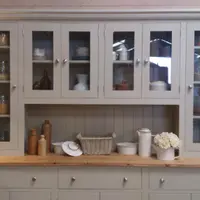 Front view of the top section with the glazed doors closed, showing the moulded cornice and tongue-and-groove back, the open shelf below holding stoneware bottles, a basket and roses