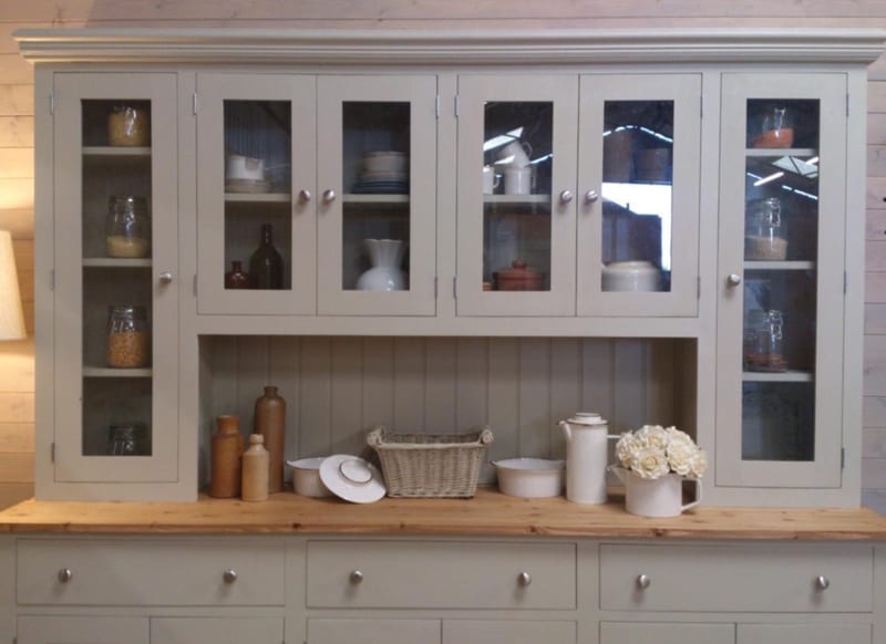 Front view of the top section with the glazed doors closed, showing the moulded cornice and tongue-and-groove back, the open shelf below holding stoneware bottles, a basket and roses