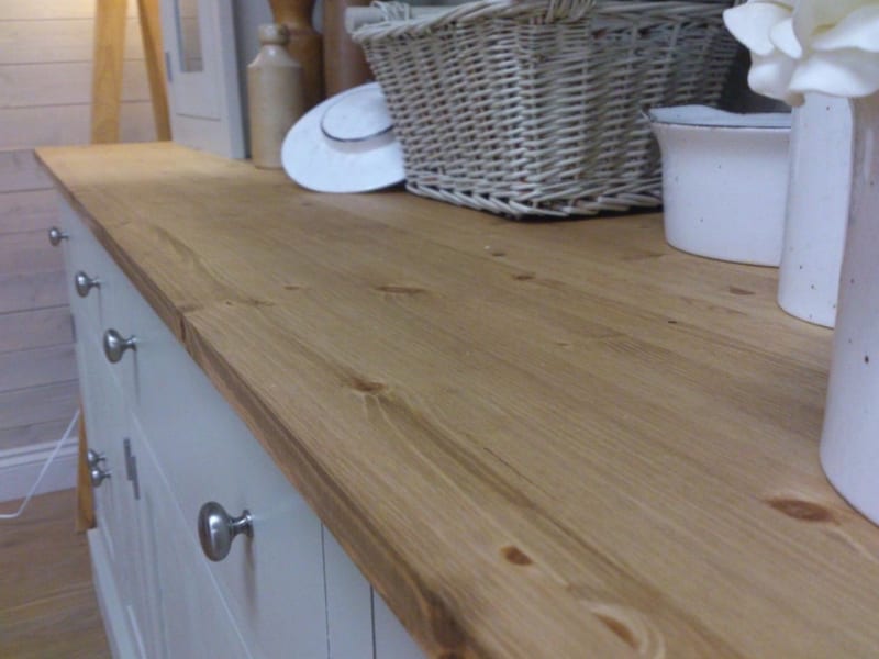 Close view along the waxed pine top, showing the knots, grain and plank joint beside a wicker basket, white enamel pot and the brushed nickel drawer knobs below