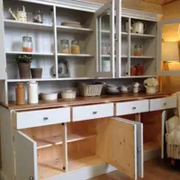 A different painted dresser with an open shelved top and glazed cupboards, its base doors open to reveal pine interiors, styled with jars, plates and a basket beside a cream armchair