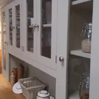 Angled view along the glazed top doors from the side, one end door open on shelves of clip-top jars, with stoneware bottles, a white basket and enamel pot on the surface below