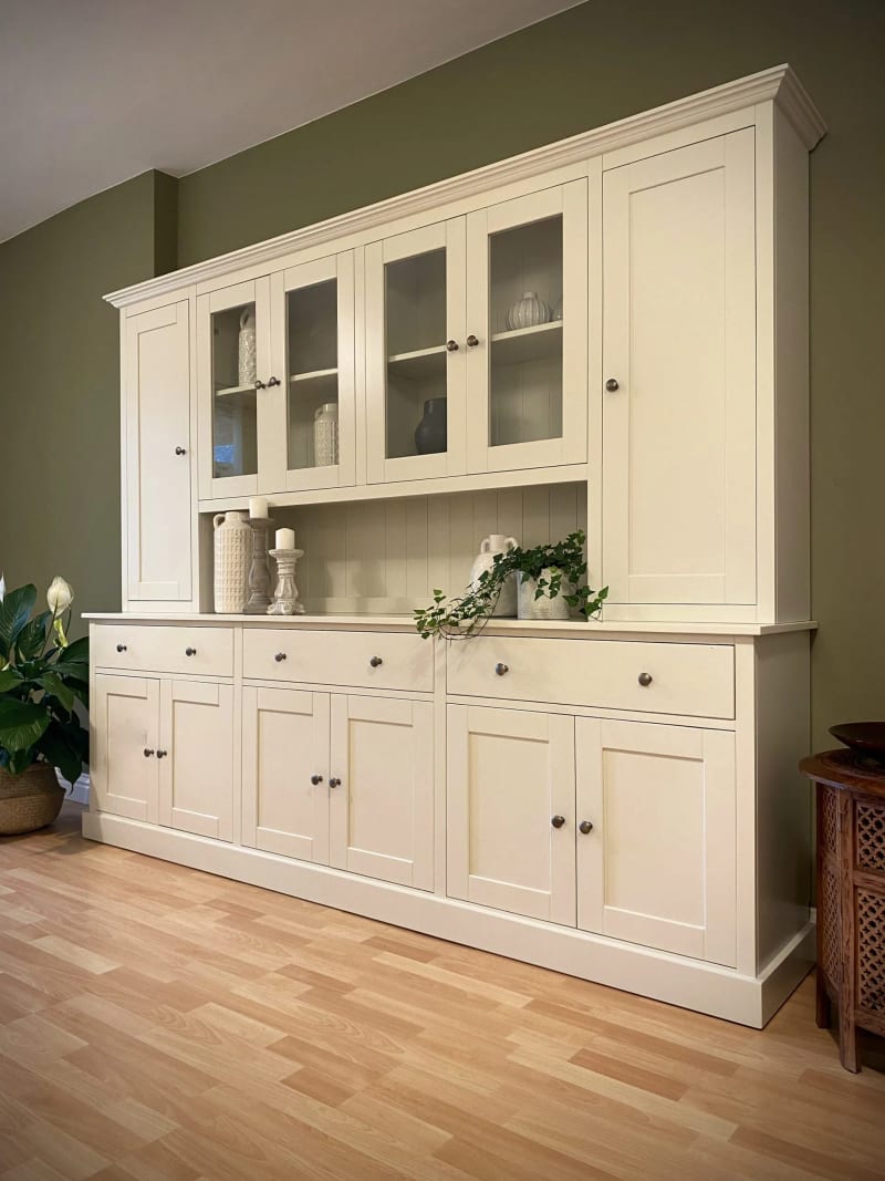 Angled view of the cream painted dresser against an olive green wall, showing four glazed doors between two tall solid end doors, with three drawers and six panelled cupboards below