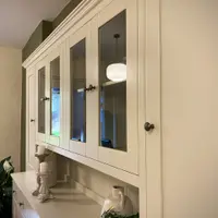 View of the upper section from the right, the glazed doors reflecting a ribbed pendant light, with the tall solid end door, moulded cornice and candlesticks on the worktop below